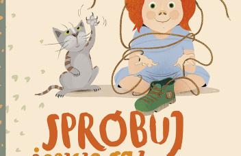 Na piaskowym tle ruda dziewczynka z kotkiem, próbująca zawiązać but